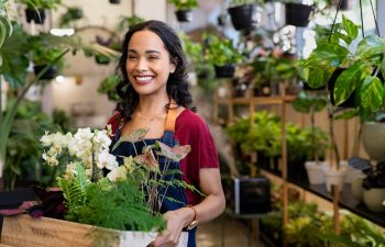 Beautiful florist holding wooden box of fresh plants at flower shop. Latin hispanic woman working in floral shop with copy space. Successful florist smiling while holding fresh flowers and plants.